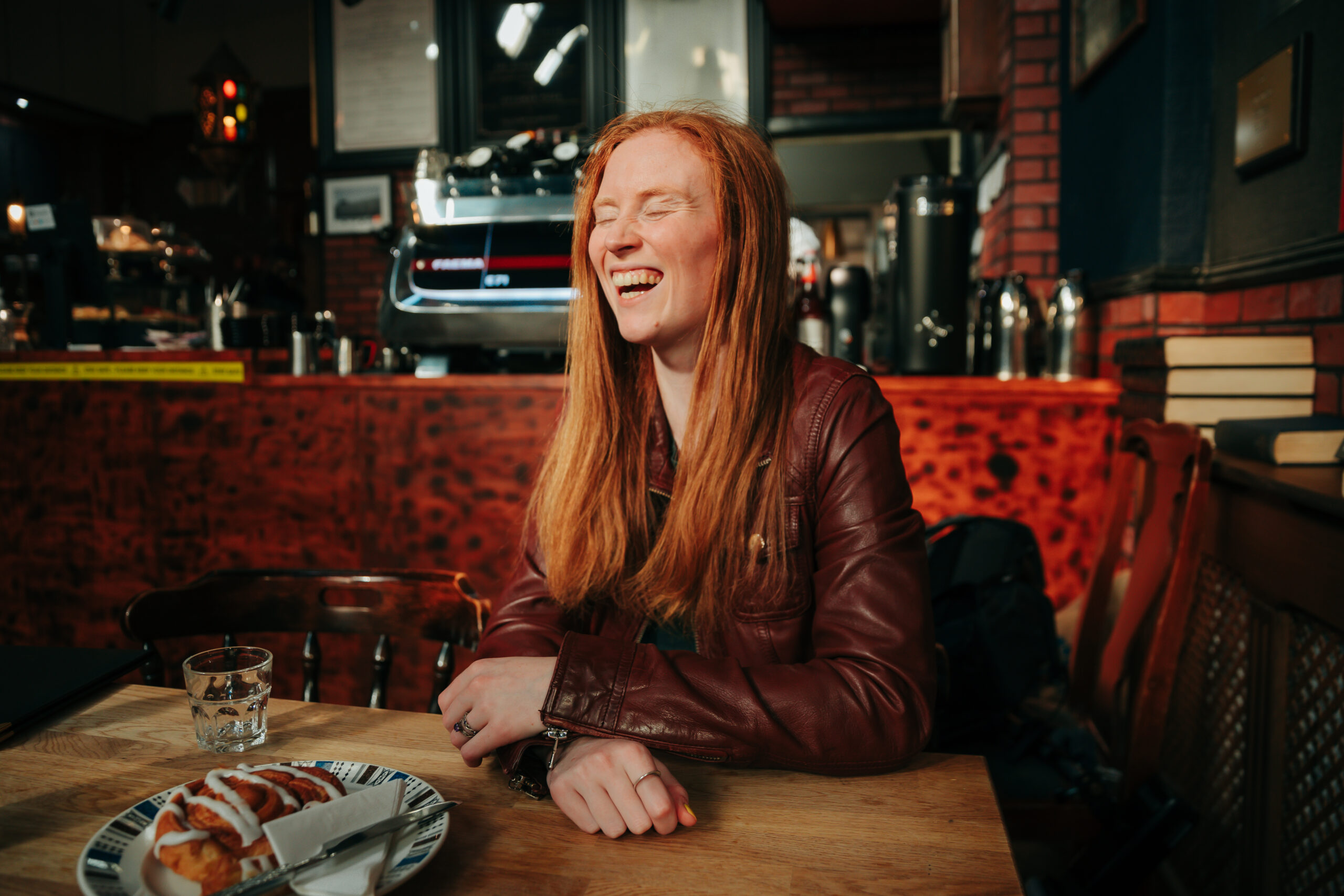 Photo of a red-haired woman sitting in a cafe laughing, with a cup of water and a pastry on the table in front of her.
