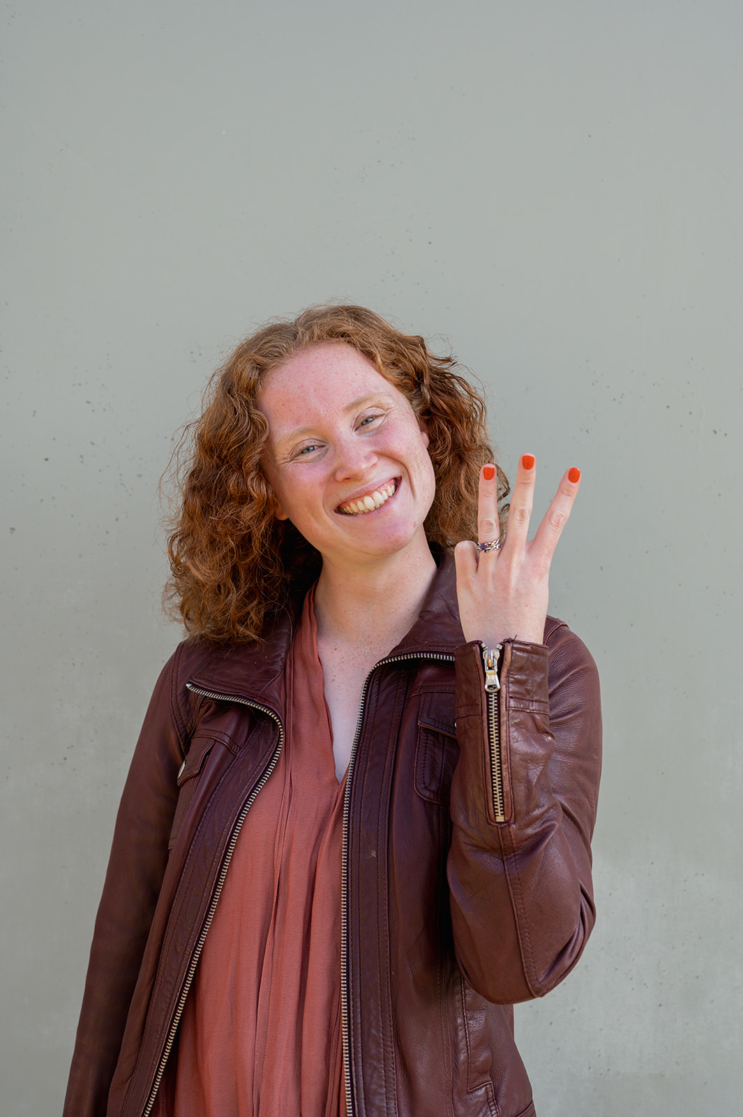 Red haired woman in a burgundy leather jacket standing against a beige concrete wall, holding 3 fingers up in the air.