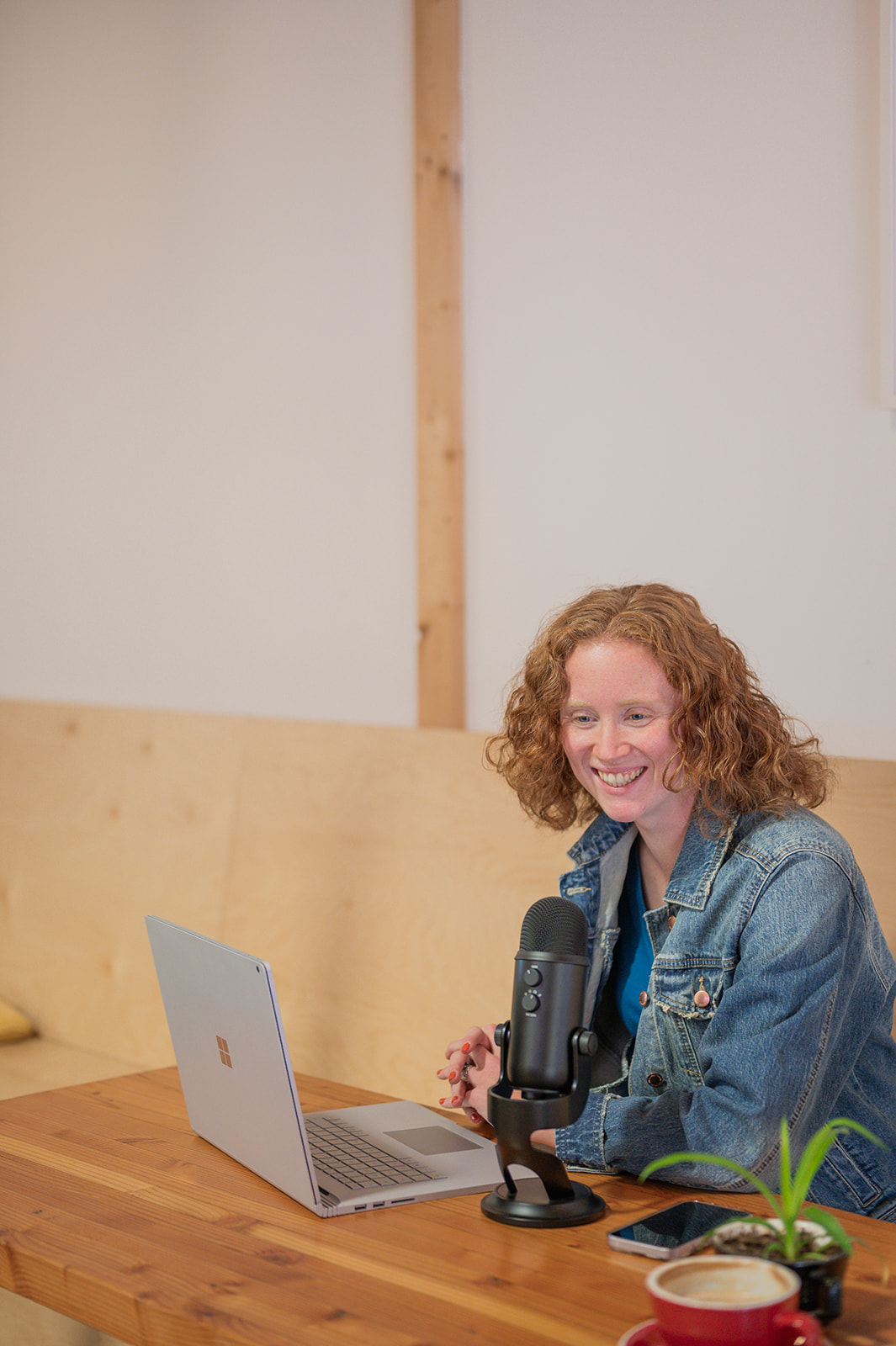 photo of a curly red haired woman sitting at a coffee shop table with her laptop and microphone.