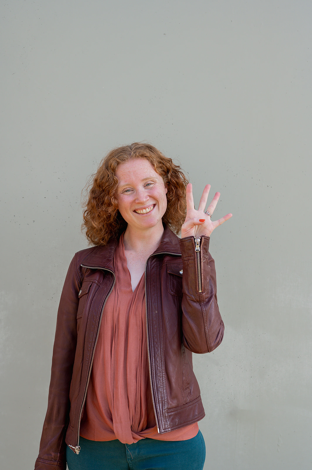 A photo of a red haired woman standing against a grey concrete wall, wearing dark teal jeans, a salmon colored shirt, and a burgandy leather jacket. She is smiling and holding up 4 fingers to the camera.