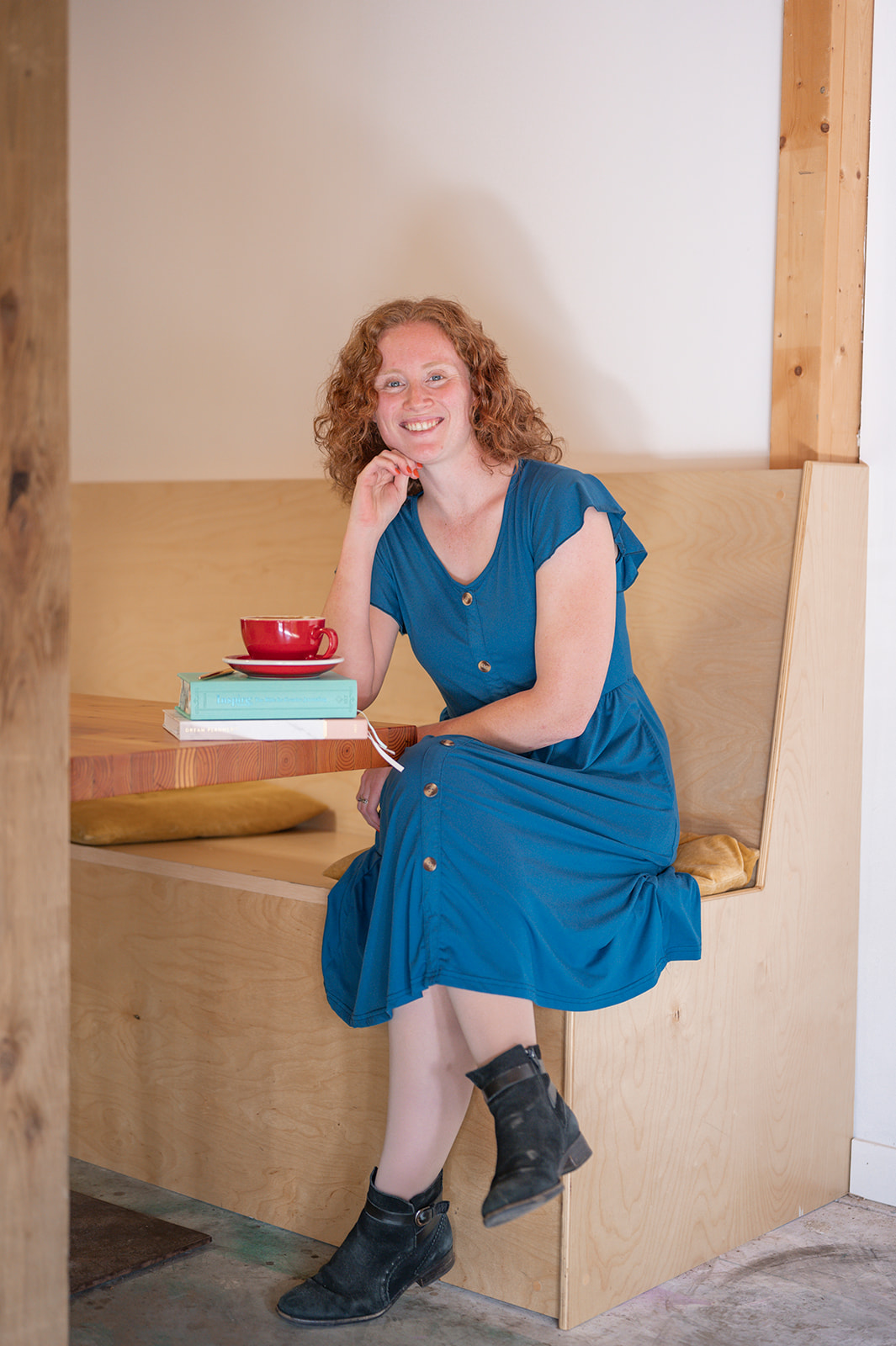 Woman sitting on a wooden bench at a coffee shop. The women has read hair and is wearing a blue dress with black anckle boots. Her coffee cup is red.