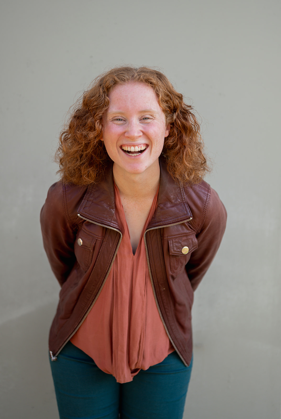 Photo of a lady with red curly hair, she is smiling and wearing a brown leather jacket with a pink blouse.